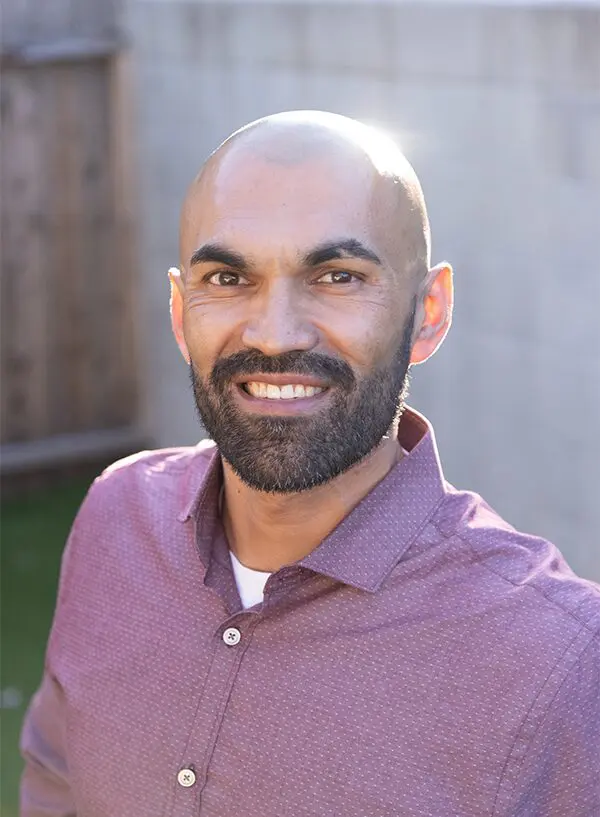 A person with a beard and shaved head smiles outdoors, wearing a maroon button-up shirt.