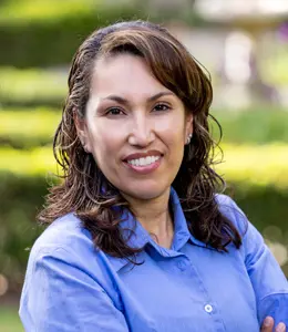 A woman with brown hair, wearing a blue shirt, smiles with arms crossed. Background shows greenery.