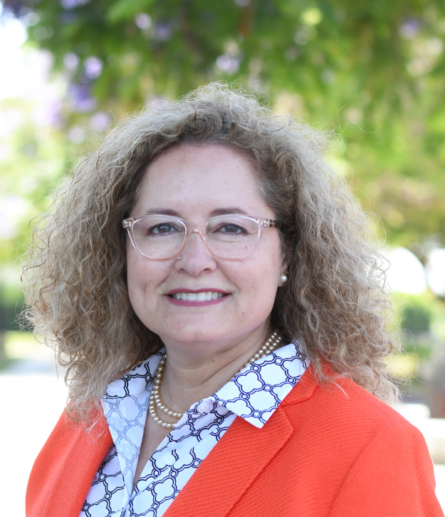 A woman with curly hair and glasses is wearing a white-patterned shirt and orange blazer, standing outdoors with greenery in the background.