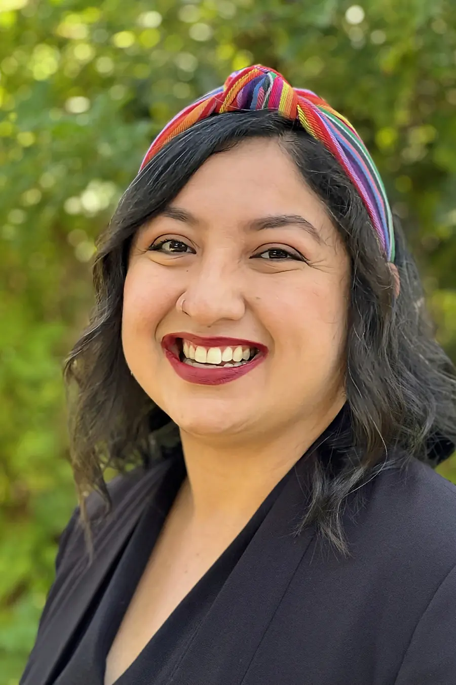 A person with dark hair and a colorful headband smiles outdoors, wearing a black jacket.