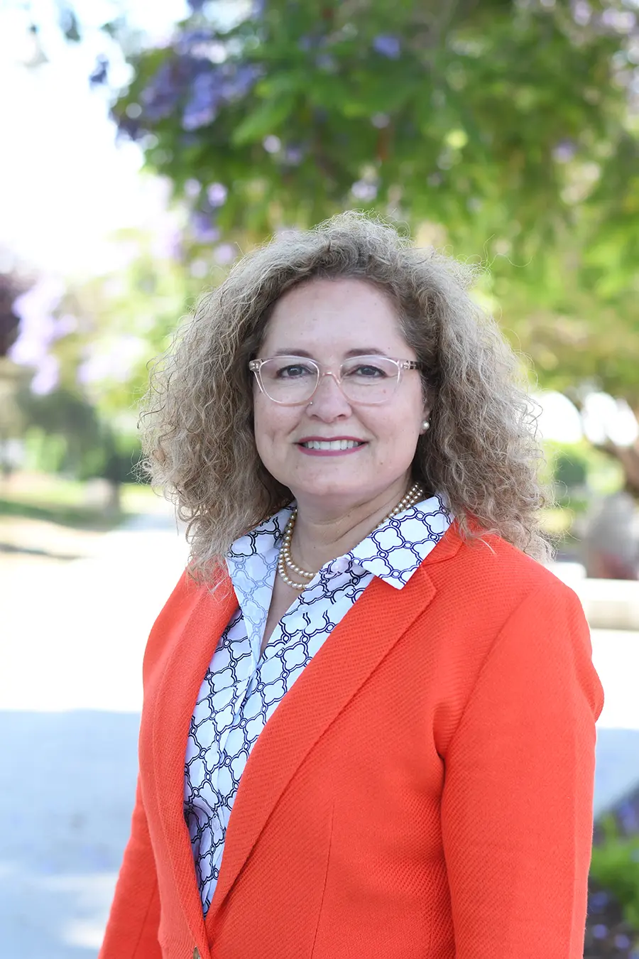 A woman with curly hair and glasses is wearing an orange blazer and patterned shirt. She stands outdoors with trees in the background, smiling at the camera.