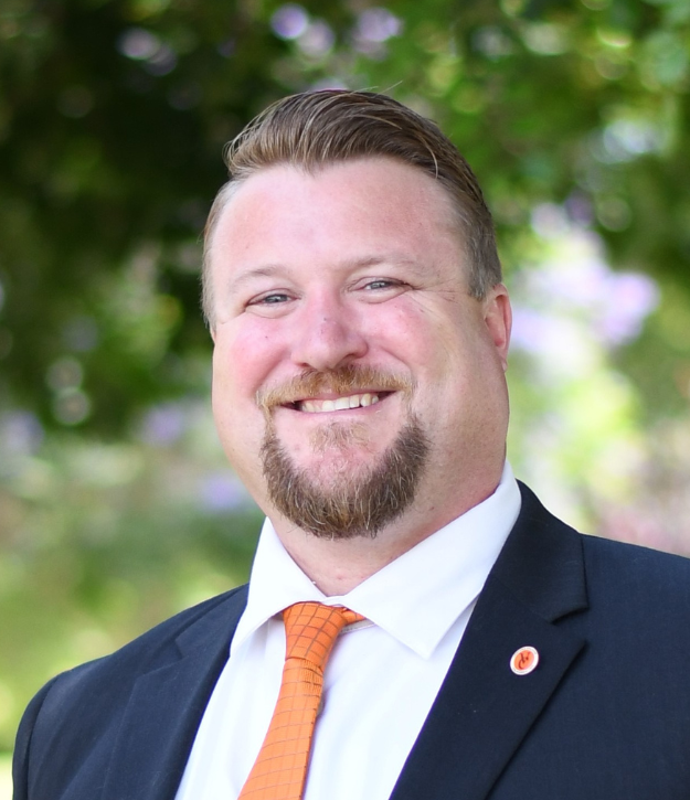 A man wearing a suit and orange tie smiles outdoors with green foliage in the background.