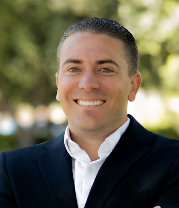 A person in a suit smiling outdoors with a blurred leafy background.