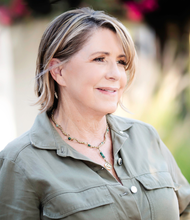 A woman with short hair and a green shirt stands outside, smiling slightly. She is wearing a necklace and there are blurred plants in the background.