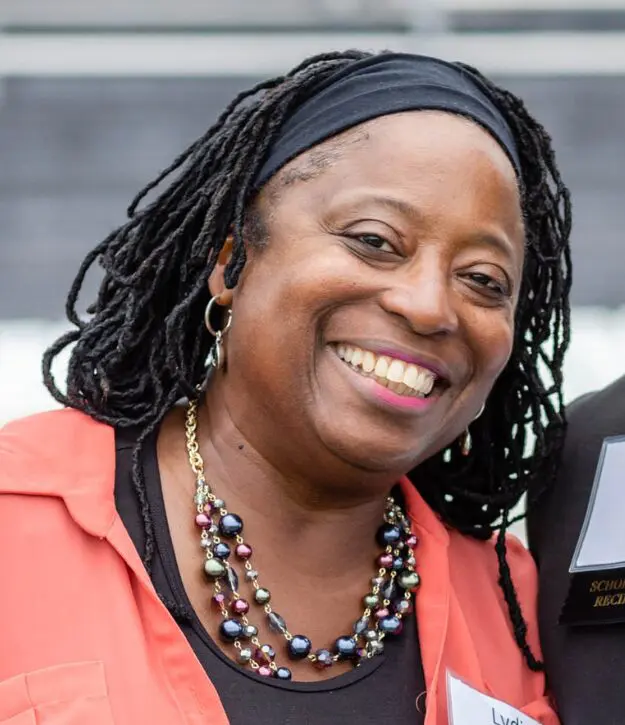 A person with braided hair and a headband smiles, wearing a colorful necklace and an orange top.