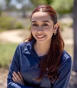 A woman with long brown hair, wearing a blue shirt, stands outdoors with arms crossed, smiling at the camera.