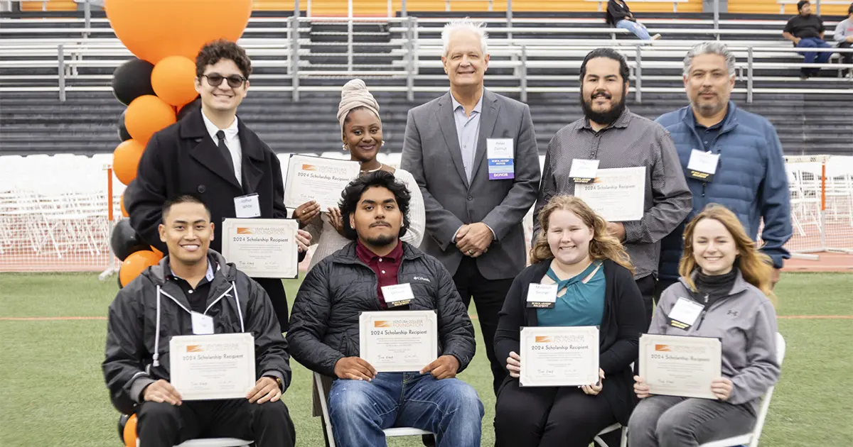 A group of people, some seated and some standing, hold certificates. They pose on a sports field with orange and black balloons in the background.