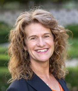 A woman with curly hair smiles, wearing a black blazer and a patterned blouse, standing outdoors with greenery in the background.