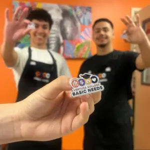Three people wearing black aprons hold Ventura College Basic Needs pins toward the camera, with a colorful painting and orange wall in the background.