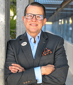 Man in a dark suit and glasses stands with arms crossed, smiling, beside a metal structure outdoors.
