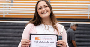 A young woman smiles while holding a Ventura College Foundation 2025 Scholarship Recipient certificate, standing outside in front of bleachers.