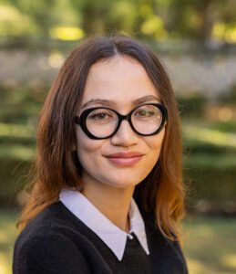 A woman with long brown hair, wearing large black glasses, a white collared shirt, and a black sweater, is standing outdoors with greenery in the background.