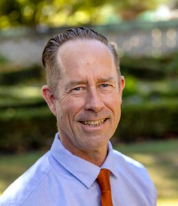 A middle-aged man with short, slicked-back hair, wearing a light blue shirt and orange tie, smiles outdoors with greenery in the background.