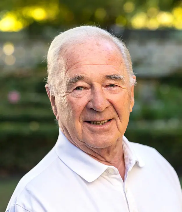 An elderly man with white hair wearing a white collared shirt smiles outdoors with greenery and a stone wall in the background.