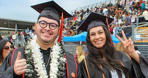 Two graduates in caps and gowns smile at the camera; one gives a thumbs up and the other makes a peace sign. A crowd is visible in the background.