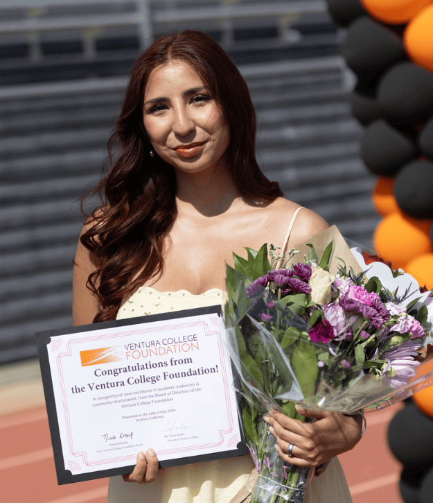 Happy graduate holding diploma and bouquet at Ventura College graduation.
