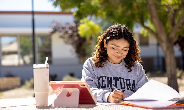 Student studying outdoors at Ventura College with books and tablet.
