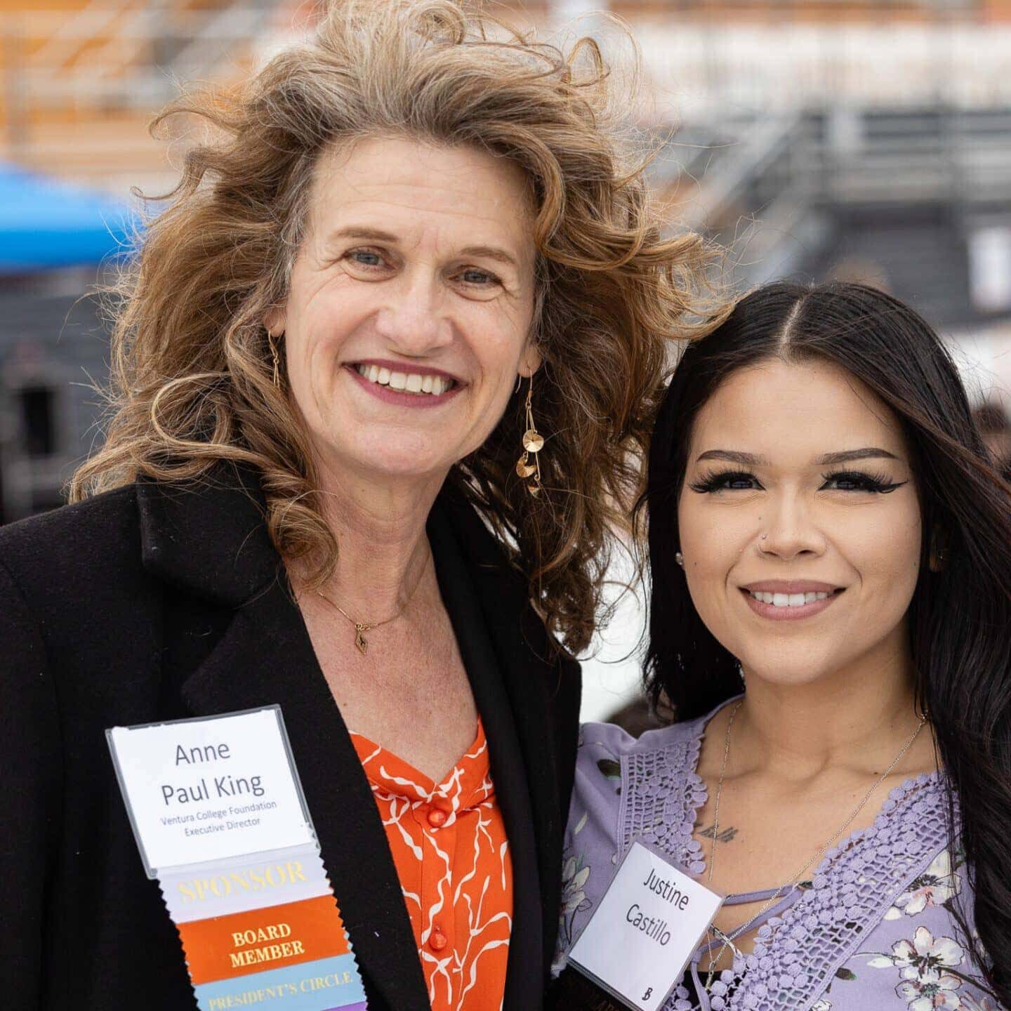 Two people are posing for a photo outdoors, both wearing name tags. The person on the left has curly hair and the person on the right has straight dark hair.